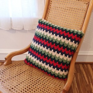 May include: A striped crocheted pillow with red, green, blue, white, and cream colored stripes. The pillow is sitting on a wicker chair.