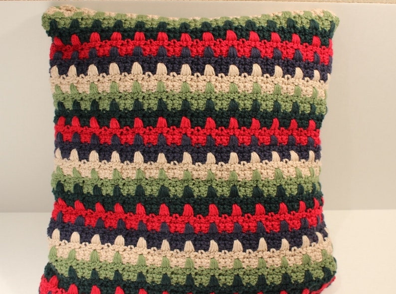 May include: A striped crocheted pillow with red, green, blue, and beige yarn. The pillow is made with a textured stitch that creates a bumpy surface.