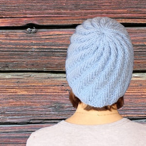 May include: A light blue knitted beanie hat with a textured pattern. The hat is worn by a person standing in front of a wooden wall.