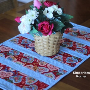 Może przedstawiać: Błękitny i czerwony bieżnik stołowy z wzorem serc. Bieżnik ozdobiony jest koszykiem z różowymi i białymi kwiatami. W prawym dolnym rogu zdjęcia widoczny jest tekst "Kimberlees Korner".