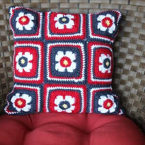 May include: A red, white, and blue crocheted pillow with a floral design. The pillow is made of yarn and has a square shape.