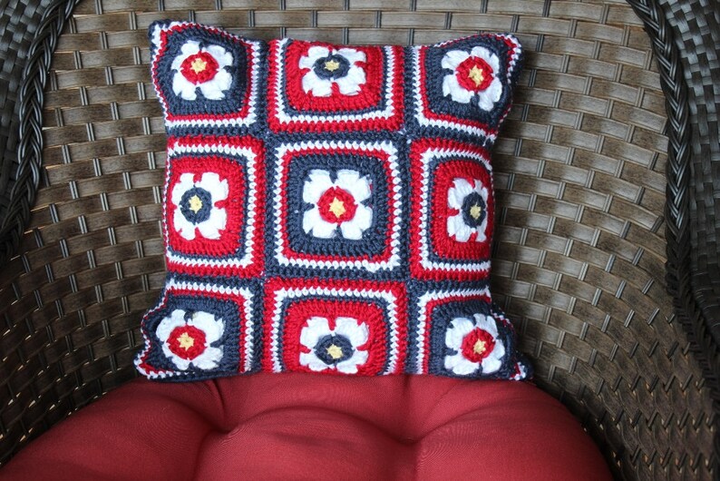 May include: A red, white, and blue crocheted pillow with a floral design. The pillow is made of yarn and has a square shape.
