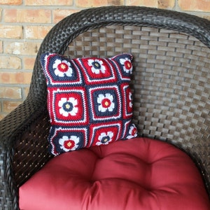 May include: A red, white, and blue crocheted pillow with a floral design sits on a brown wicker chair with a red cushion.