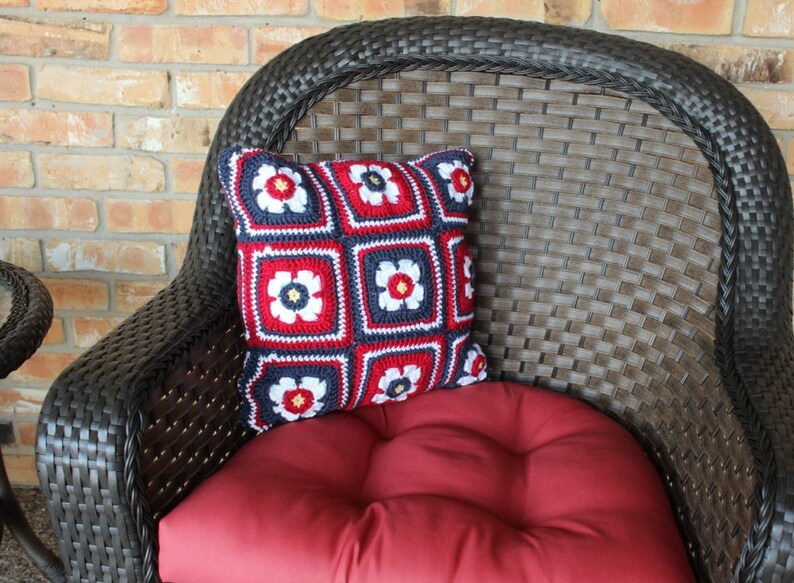 May include: A red, white, and blue crocheted pillow with a floral design sits on a brown wicker chair with a red cushion.