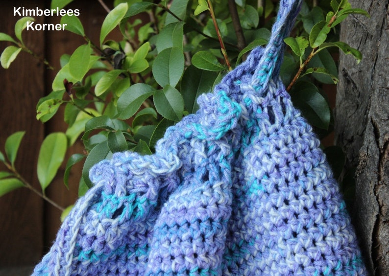 Puede incluir: Bolso de ganchillo hecho a mano en tonos azules y morados, con cierre de cord&oacute;n y un patr&oacute;n de puntada texturizada. El bolso cuelga de una rama de &aacute;rbol, con el texto "Kimberlees Korner" visible.