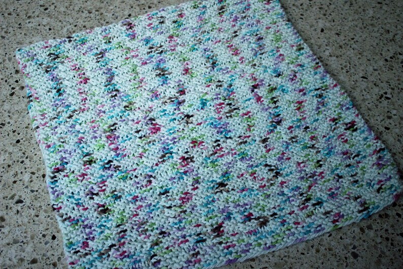 May include: A white knitted dishcloth with a multi-colored yarn pattern. The yarn is a mix of pink, blue, green, brown, and white.