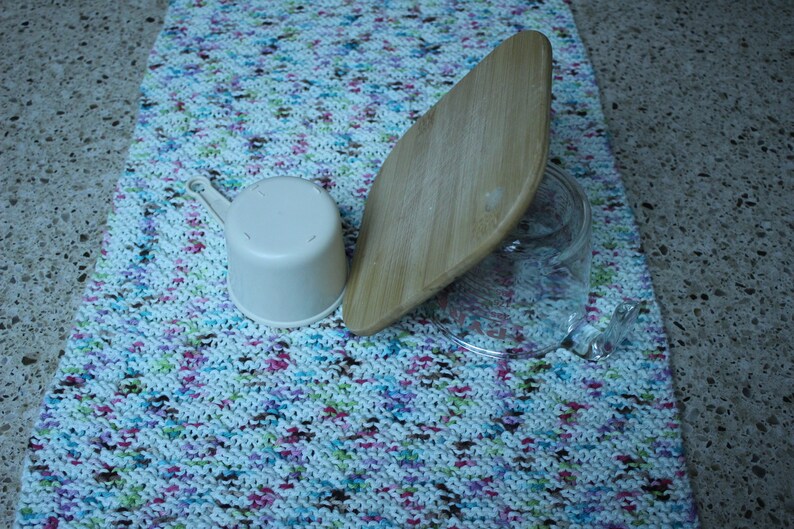 May include: A white plastic measuring cup, a wooden cutting board, and a clear glass jar on a colorful knitted kitchen towel.