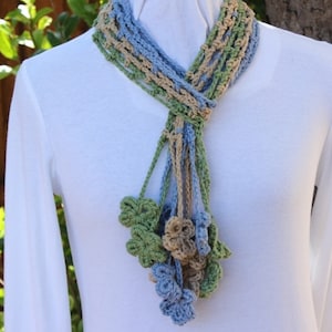 May include: A crocheted scarf with a blue, green, and beige color scheme. The scarf has a long fringe with multiple flower-shaped embellishments.