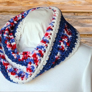 Op de afbeelding: Een witte, rode en blauwe gecrochete oneindige sjaal met een glinsterend draad dat door de hele sjaal is geweven.