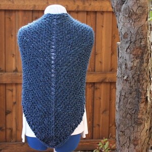 May include: A dark blue knitted shawl with a textured pattern. The shawl is displayed on a mannequin, showcasing its triangular shape and open back design. The background features a wooden fence and a tree trunk.