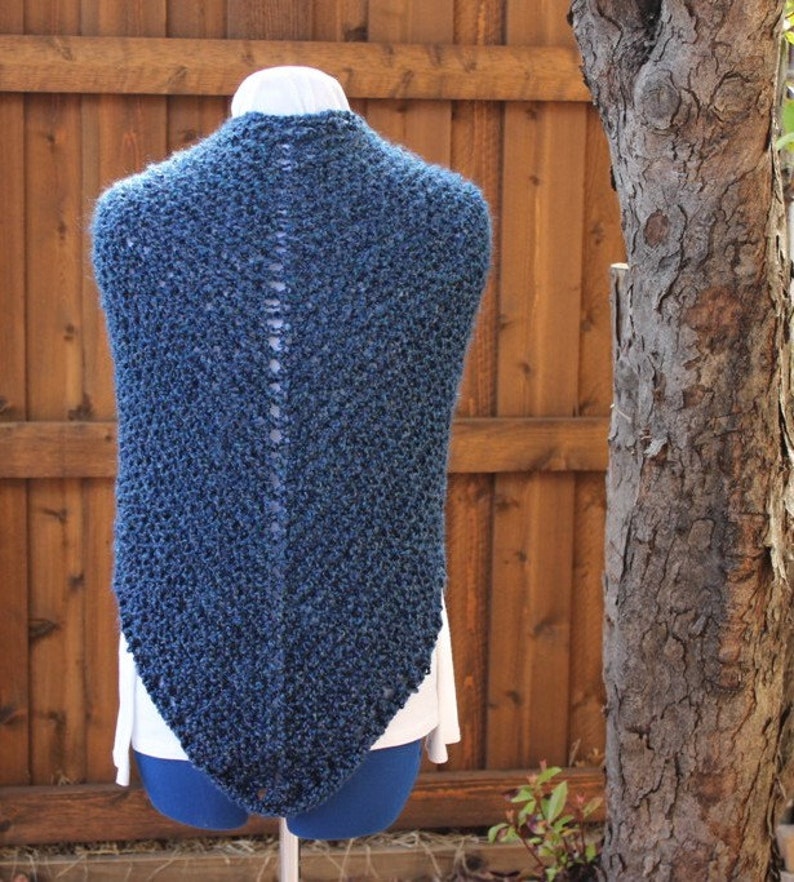 May include: A dark blue knitted shawl with a textured pattern. The shawl is displayed on a mannequin, showcasing its triangular shape and open back design. The background features a wooden fence and a tree trunk.