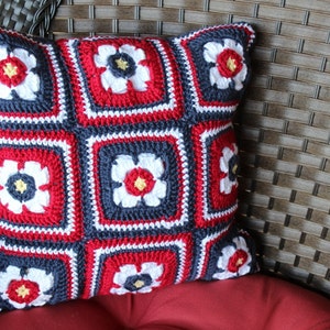 May include: A crocheted pillow with a red, white, and blue floral pattern. The pillow is made of yarn and has a square shape.