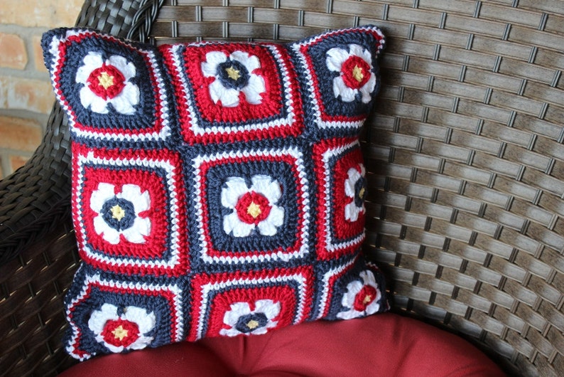 May include: A crocheted pillow with a red, white, and blue floral pattern. The pillow is made of yarn and has a square shape.