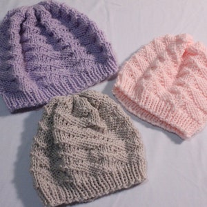 May include: Three knitted hats in different colors: purple, gray, and pink. The hats are made with a textured stitch pattern.