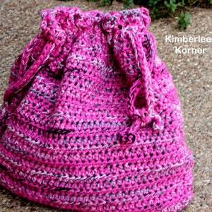 Puede incluir: Un bolso de ganchillo hecho a mano con cord&oacute;n en tonos rosa, blanco y gris. El bolso presenta un patr&oacute;n de puntadas texturizadas y un cierre de cord&oacute;n trenzado. El texto "Kimberlees Korner" es visible en el lateral del bolso.
