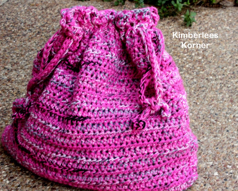 Puede incluir: Un bolso de ganchillo hecho a mano con cord&oacute;n en tonos rosa, blanco y gris. El bolso presenta un patr&oacute;n de puntadas texturizadas y un cierre de cord&oacute;n trenzado. El texto "Kimberlees Korner" es visible en el lateral del bolso.