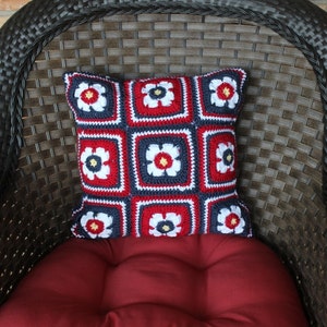 May include: A red, white, and blue crocheted pillow with a floral design. The pillow is sitting on a brown wicker chair.