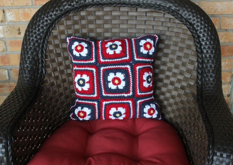 May include: A red, white, and blue crocheted pillow with a floral design. The pillow is sitting on a brown wicker chair.