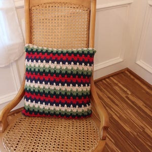 May include: A striped crocheted pillow with red, green, white, and blue stripes. The pillow is sitting on a wooden chair with a woven cane seat.