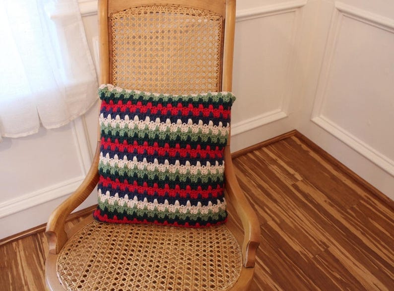 May include: A striped crocheted pillow with red, green, white, and blue stripes. The pillow is sitting on a wooden chair with a woven cane seat.