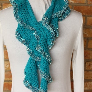 May include: A teal blue crocheted scarf with a ruffled edge. The scarf is made with a sparkly yarn and is draped around a white shirt.