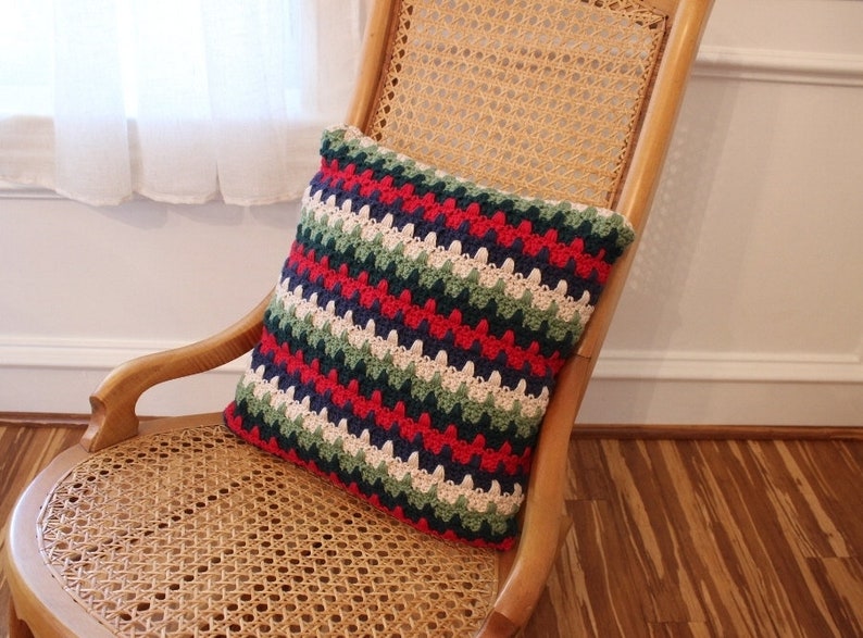 May include: A striped crocheted pillow with red, green, blue, white, and cream colored stripes. The pillow is sitting on a wicker chair.