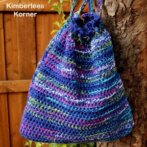 Puede incluir: Un bolso de ganchillo azul hecho a mano con cord&oacute;n y detalles coloridos en rosa, verde y blanco. El bolso cuelga de un &aacute;rbol y presenta el texto "Kimberlees Korner" en la parte superior izquierda.