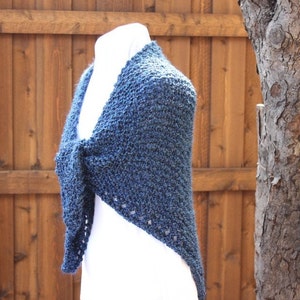 May include: A blue knitted shawl draped over a white form. The shawl is a triangular shape with a textured knit pattern. The color is a deep blue with subtle variations, suggesting a hand-dyed or variegated yarn. The shawl is knotted at the front.