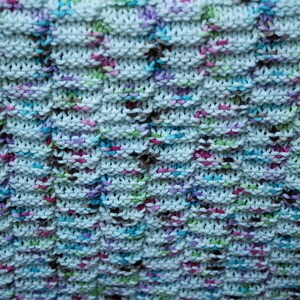 May include: A close-up of a knitted dishcloth with a white background and multi-colored yarn. The yarn is in shades of blue, green, pink, and brown.
