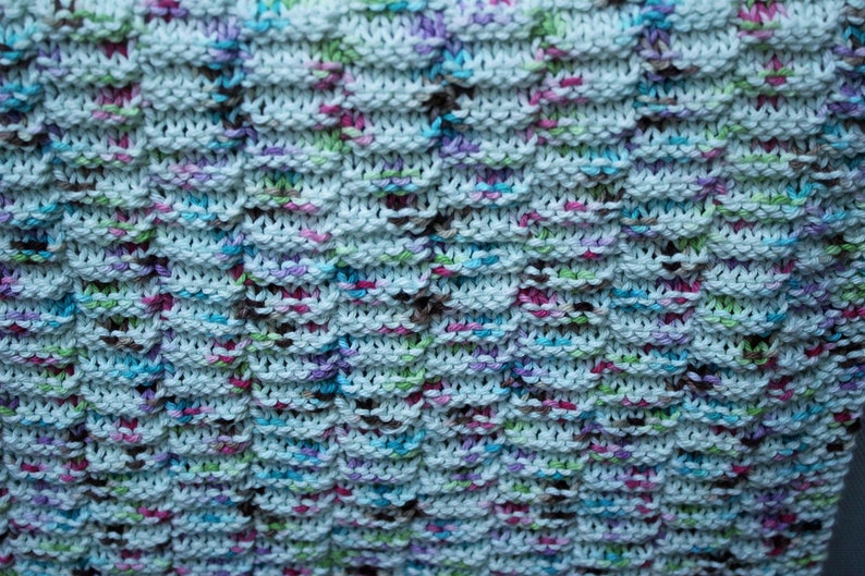 May include: A close-up of a knitted dishcloth with a white background and multi-colored yarn. The yarn is in shades of blue, green, pink, and brown.