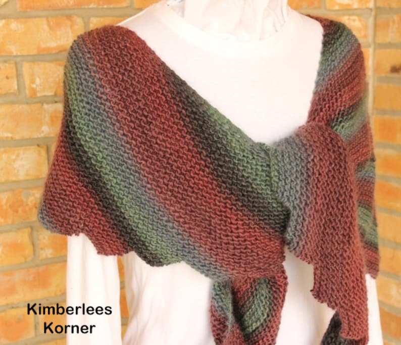 May include: A hand-knitted shawl in shades of red, green, and gray, draped over a white top. The shawl features a textured stitch pattern and a scalloped edge. The text "Kimberlees Korner" is visible in the lower left corner.