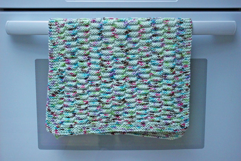 May include: A colorful knitted dishcloth with a textured pattern hangs on a white oven door.