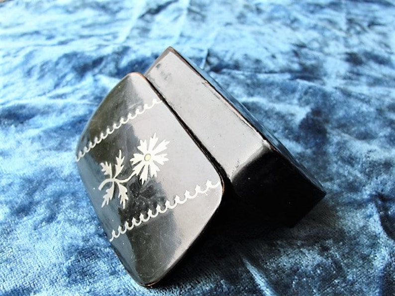 May include: A small black box with a silver floral design on the lid. The box is closed and the lid is slightly ajar.