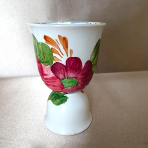 May include: White ceramic egg cup with a floral design. The cup features a large, deep red flower with green and orange accents. The base is a bell shape. The rim has a blue and white pattern.