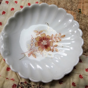 May include: A white porcelain dish with a scalloped edge and a floral design in shades of brown and gold. The dish is ideal for serving small snacks or desserts.