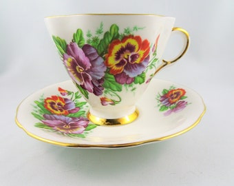 May include: Cream-colored teacup and saucer set with a floral pansy design in shades of purple, orange, and yellow. The cup and saucer have gold trim. The teacup has a curved handle and sits on a small foot.