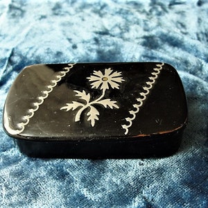 May include: A small black rectangular box with a silver floral design. The box has a silver border and a silver flower with a white center.
