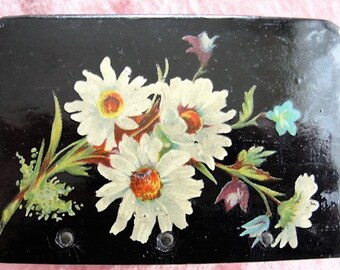 May include: A black box with a floral design painted on the top. The flowers are white daisies with yellow centers and green leaves. There are also blue and purple flowers in the design.