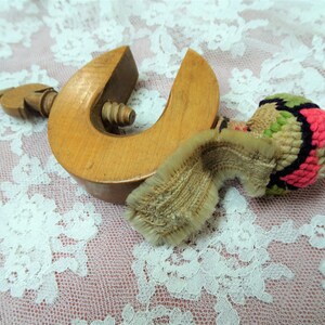 Könnte beinhalten: Eine Holzklemme mit einer Schraube und einer kleinen, bunten, gehäkelten Kugel daran. Die Klemme ist aus hellem Holz und hat eine natürliche Oberfläche. Die Kugel ist aus rosa, grünem und schwarzem Garn.