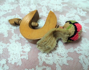 May include: A wooden clamp with a screw and a small, colorful, crocheted ball attached to it. The clamp is made of light wood and has a natural finish. The ball is made of pink, green, and black yarn.
