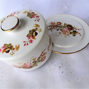 May include: A white ceramic cake plate with a gold trim and a floral design. The plate has a matching lid with a knob on top.