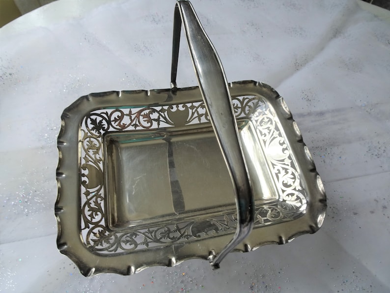 K&ouml;nnte beinhalten: Silberfarbener rechteckiger Korb mit Henkel. Der Korb hat ein dekoratives, durchbrochenes Design an den R&auml;ndern und einen gewellten Rand. Der Griff ist gew&ouml;lbt und an den Seiten befestigt. Ein Vintage-Servierst&uuml;ck.