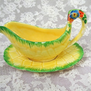 Pode incluir: Um barco de molho de cerâmica amarelo com um design de padrão verde e amarelo. A alça tem a forma de uma folha e tem flores pintadas em vermelho, azul e amarelo. O barco está sentado em um pires correspondente.