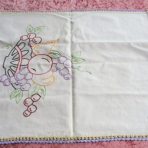 May include: White embroidered table runner with a crocheted border. The embroidery features a basket of fruit with grapes, pears, and apples in shades of purple, yellow, red, and green.