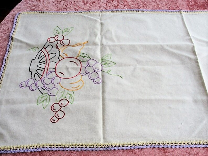 May include: White embroidered table runner with a crocheted border. The embroidery features a basket of fruit with grapes, pears, and apples in shades of purple, yellow, red, and green.