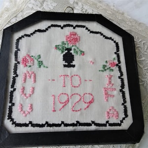 以下が含まれることがあります： ピンクのバラと緑の葉が刺繍された額装されたクロスステッチのサンプル。サンプルには「To - 1929」と書いてあります。