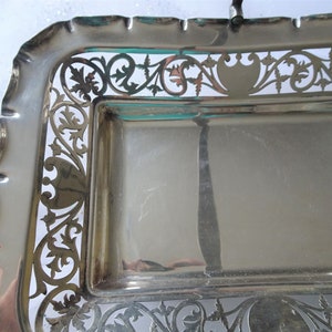K&ouml;nnte beinhalten: Ein rechteckiges Serviertablett in Silberfarbe mit kunstvollen, durchbrochenen floralen und scrollartigen Designs an den R&auml;ndern. Das Tablett hat einen gewellten Rand und einen Griff. Die Oberfl&auml;che ist reflektierend.