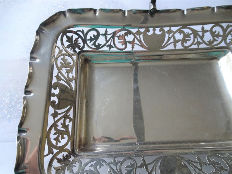 K&ouml;nnte beinhalten: Ein rechteckiges Serviertablett in Silberfarbe mit kunstvollen, durchbrochenen floralen und scrollartigen Designs an den R&auml;ndern. Das Tablett hat einen gewellten Rand und einen Griff. Die Oberfl&auml;che ist reflektierend.