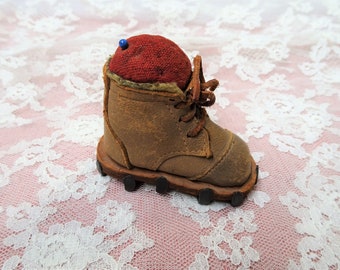 May include: A vintage leather pincushion shaped like a boot with a red fabric top and a blue pin. The boot has a lace-up closure and a studded sole.