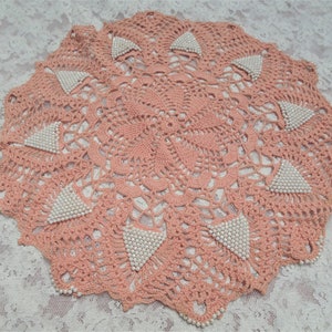 May include: A pink crocheted doily with a delicate floral pattern and white pearl accents.